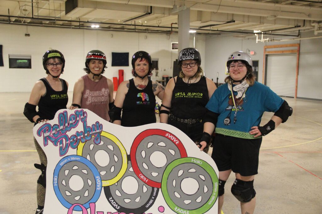 Ursa Minor coaches Comma Chameleon Head Coach Weird Val, Helly Bean, Killer Cuddy and SlayStation2 standing behind sign with text Roller Derby and a painting of five different colored roller derby wheels. 