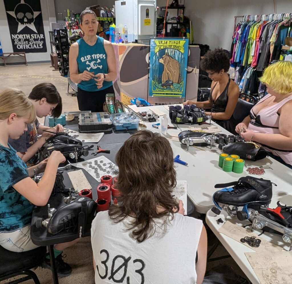 A coach talks leads a gear maintenance clinic for skaters. Five teenaged skaters are listening and working on their skates.
